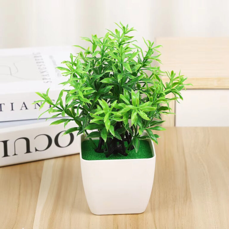 Mini artificial green plant in a modern white pot sitting on a wooden desk next to books.